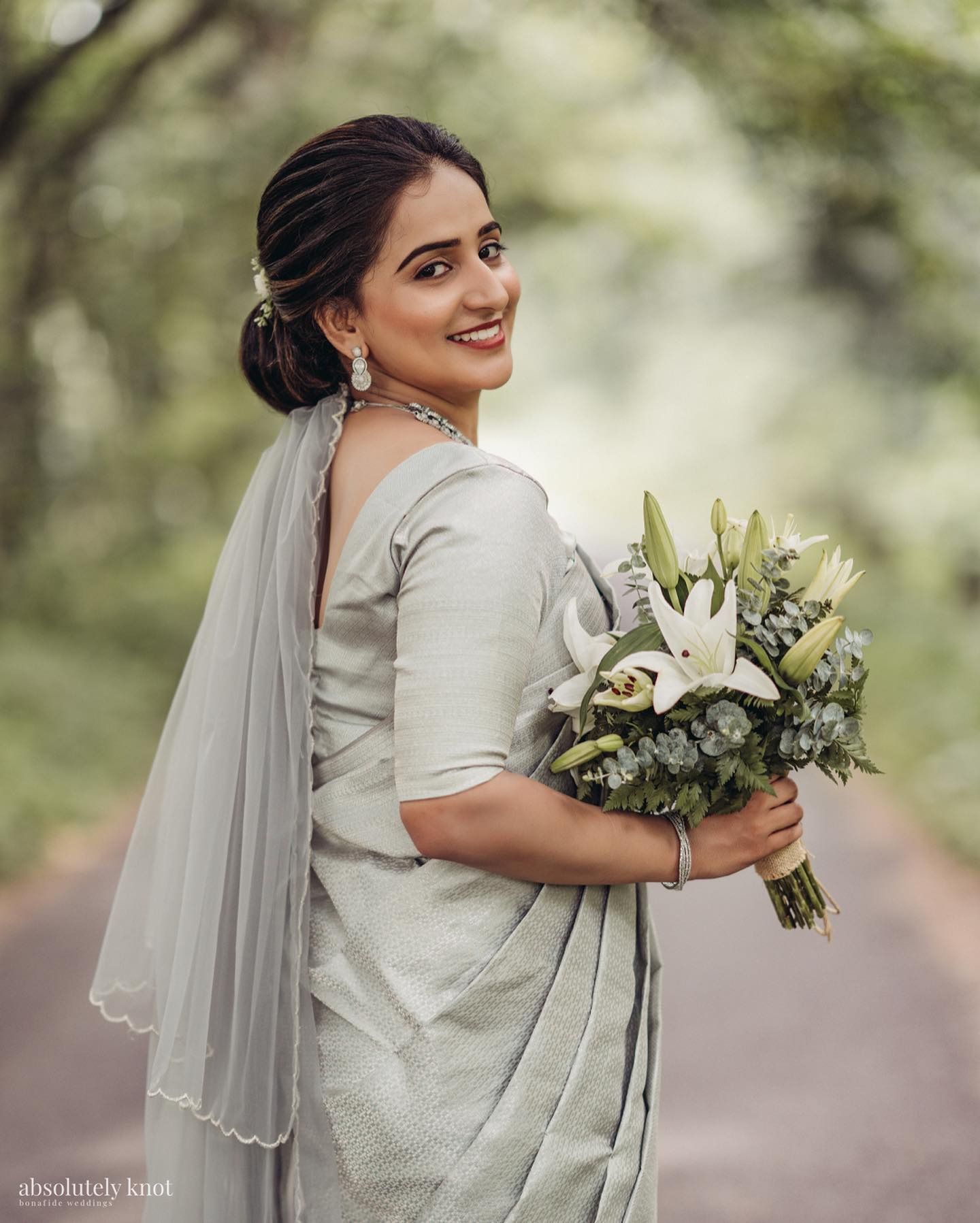 A bride in an ivory saree and veil looking back over her shoulder on a tree-lined path
