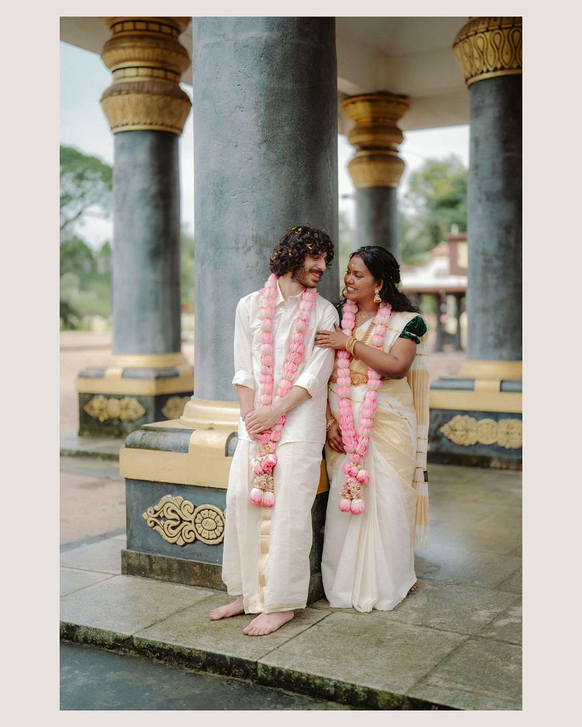 A couple wearing wedding garlands, standing between temple pillars